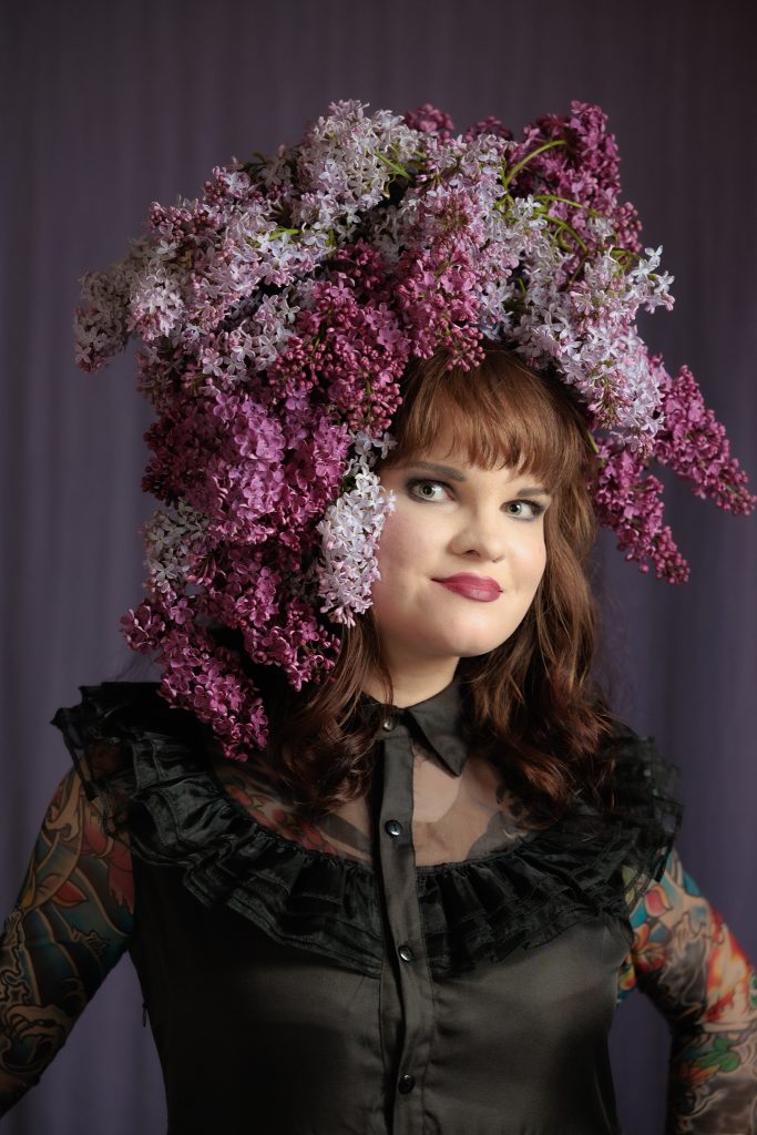 Arina Bazarbajeva melnā terpā uz tumši violeta fona ar ceriņu galvas rotu pozē fotogrāfam.