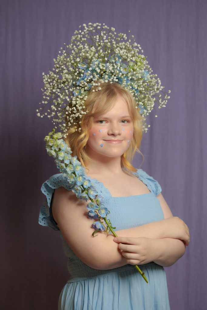 Emīlija Liepiņa gaiši zilā kleitā ar neaizmirstulīšu vainagu galvā un gaiši zilu garu ziedu rokās pozē fotogrāfam uz violeta fona.