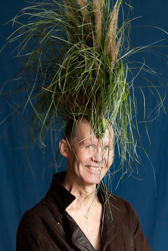 Ritvars Brālis tumšā jakā smaidot, pozē fotogrāfam ar lielu tumšu zaļu niedru un zeltainu smilgu vainagu galvā, kas atgādina pnaka grebeni.